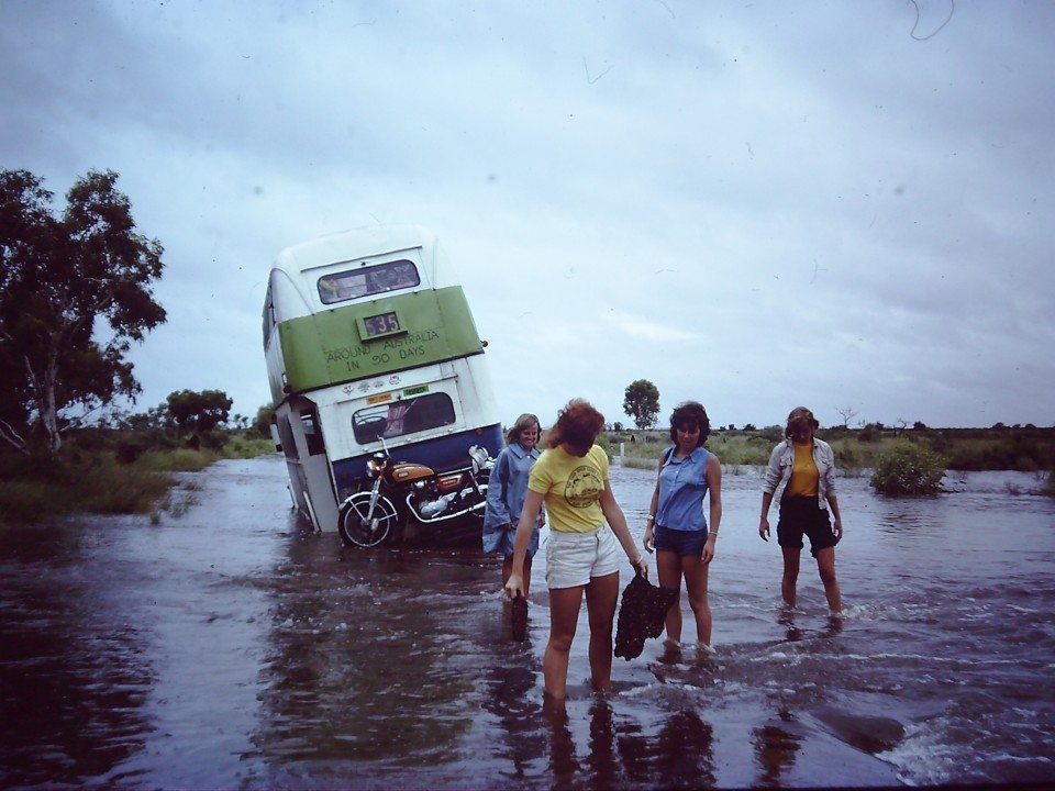 The double-decker bus tilted in floodwater, three people wading beside it