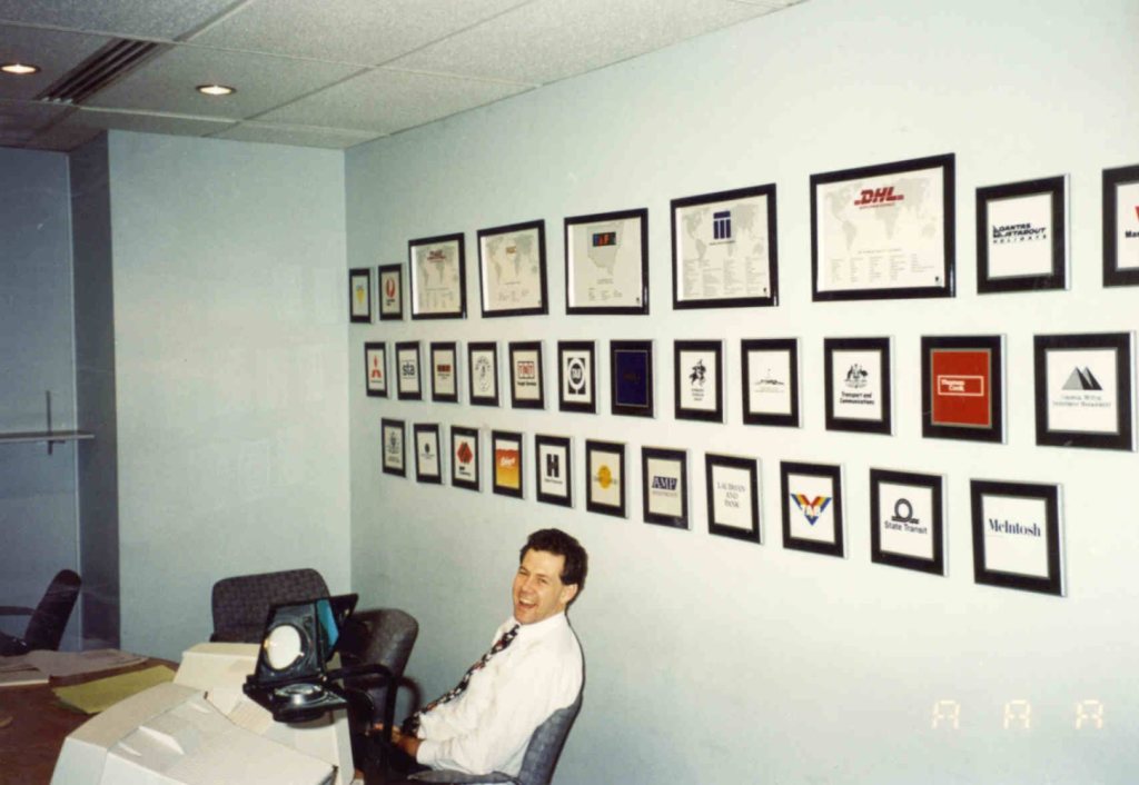 Guy at a desk, laughing, beside a wall of framed client logos